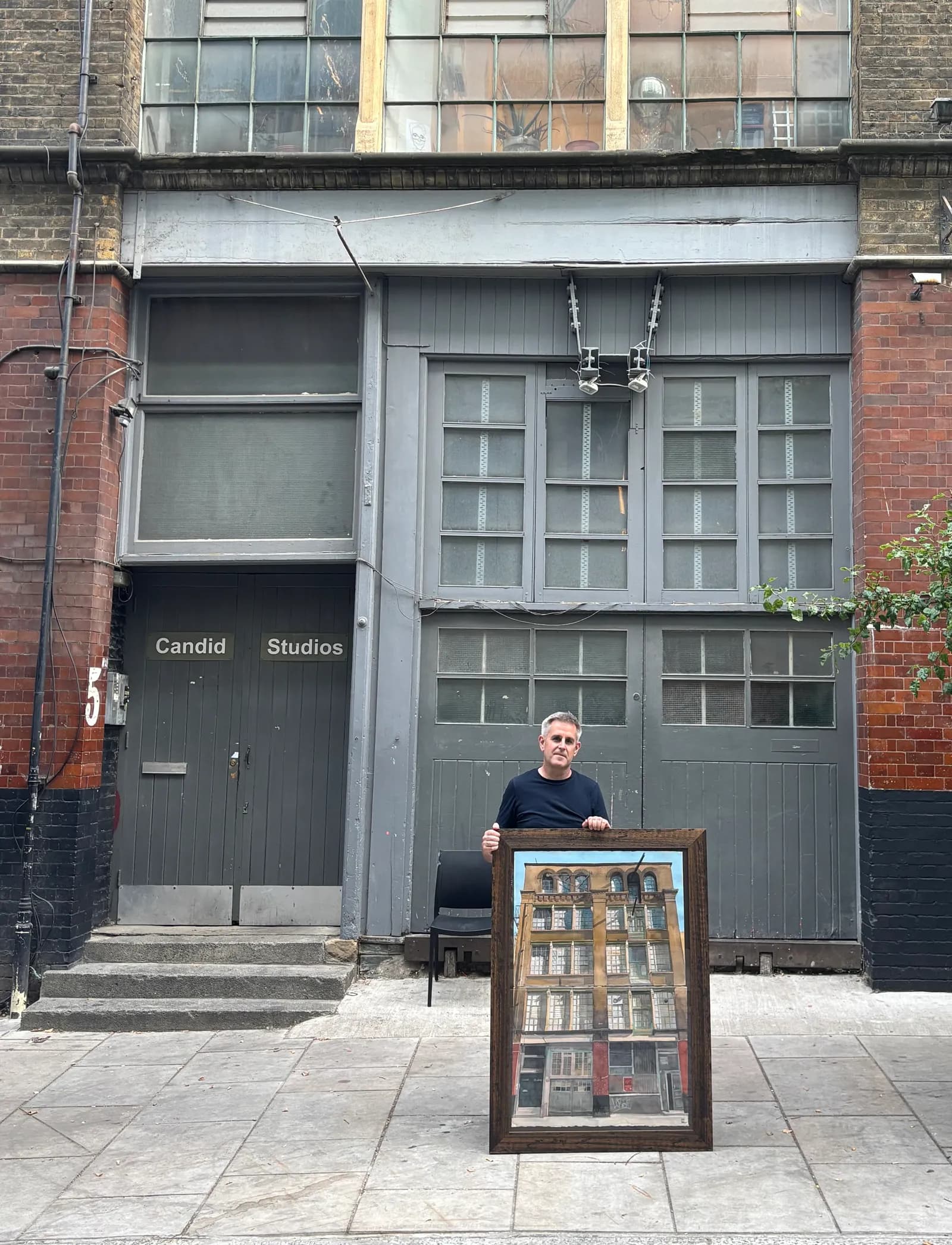 A man in a black t-shirt stands outside a gray building with a painting of a multi-story building in a wooden frame in front of him.