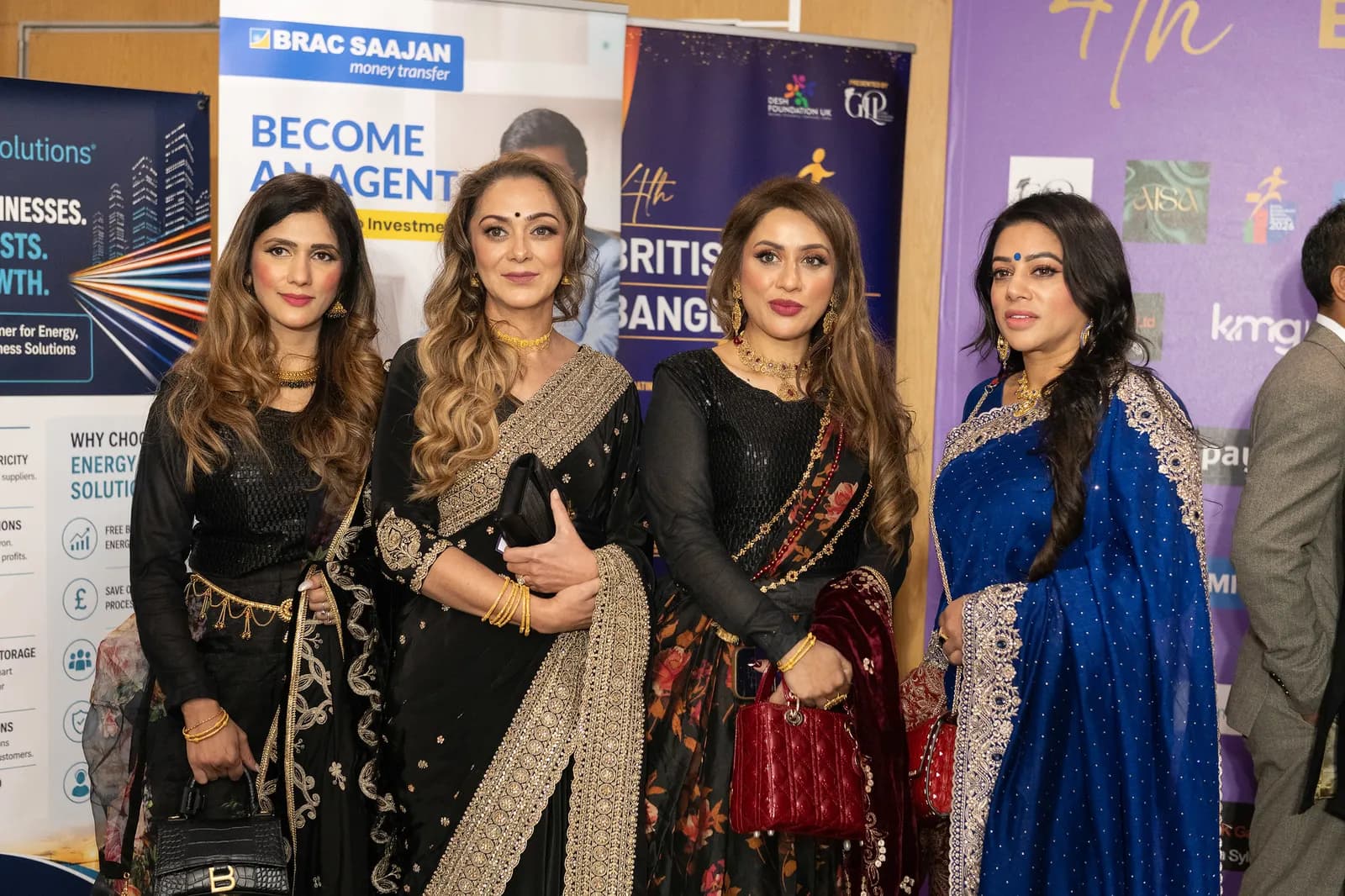 Four women in traditional Indian attire pose for a photo at an event, with three wearing black saris and one in a royal blue sari.