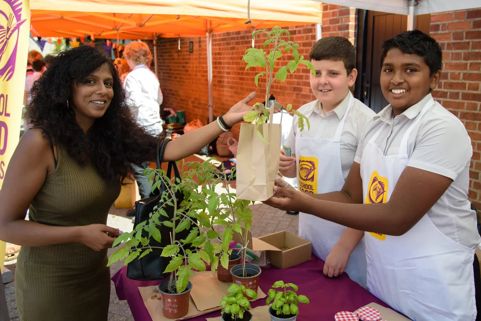A smiling woman with long dark curly hair reaches for a potted plant in a brown paper bag held by two young boys wearing white aprons, behind a table with more plants.